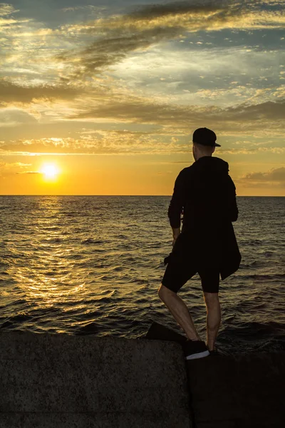 Un joven está de pie junto al mar y mirando el atardecer — Foto de Stock