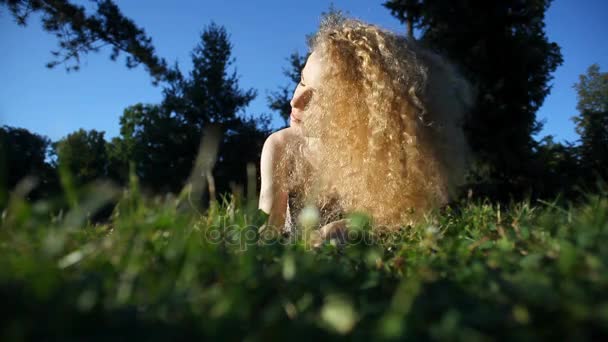 Mulher bonita com cabelo longo encaracolado louro chicoteando seu cabelo e apreciando a luz do sol deitado na grama ao ar livre cercado pela natureza Vídeo De Stock