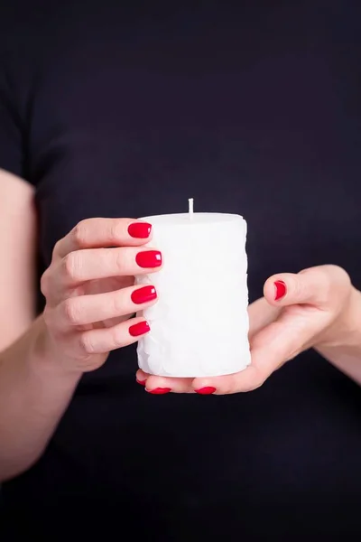 Beautiful female hands holding white candle with flower pattern. Manicure with red nail polish. Black background — Stockfoto