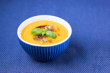 Kabak tohumlu ev yapımı balkabağı kreması çorbası, kabak yağı, fesleğen yaprağı ve mavi zemin üzerinde zerdeçal.