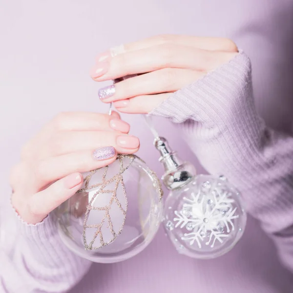 Beautiful female hands holding two transparent Christmas balls on the pale violet background. Manicure with pink color nail polish with shiny design, nude manicure — 스톡 사진