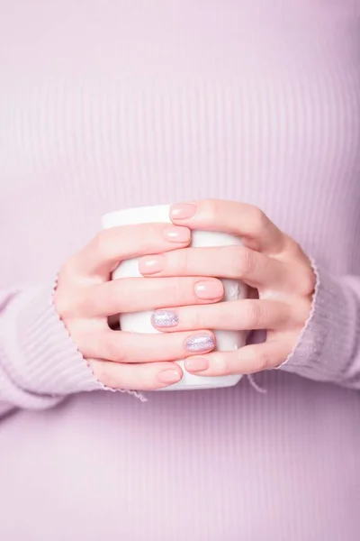 Schöne weibliche Hände, die weiße Tasse auf dem blassvioletten Hintergrund halten. Maniküre mit pinker Farbe Nagellack mit glänzendem Design, Nude-Maniküre. Kopierraum — Stockfoto