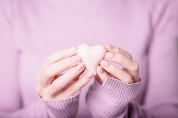 Soluk menekşe pembe arka planda pembe kalp tutan güzel kadın elleri. Pembe ojeli manikür, eğrelti otuyla çıplak manikür.
