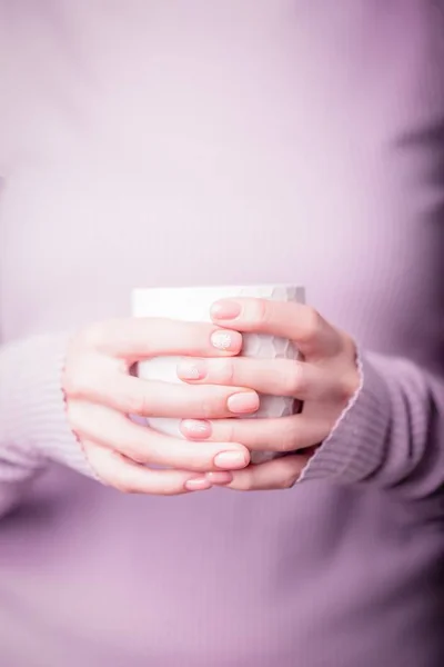 Vackra kvinnliga händer som håller vit kopp på den bleka violetta bakgrunden. Manikyr med rosa färg nagellack. Naken manikyr med ormbunke och monstera mönster. Kopiera utrymme — Stockfoto