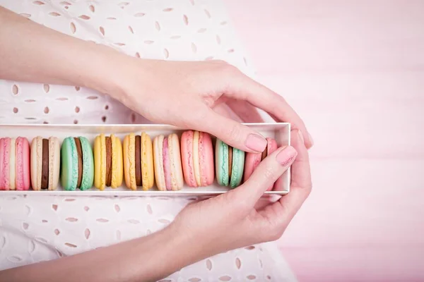 Hermosas manos femeninas sosteniendo caja de regalo de deliciosos macarrones de colores franceses sobre fondo rosa. Vista superior, espacio de copia —  Fotos de Stock