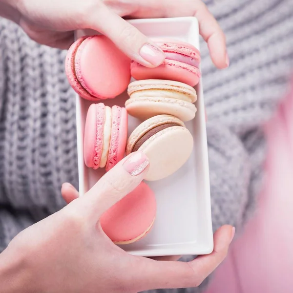 Mains féminines tenant une assiette rectangulaire blanche avec de délicieux macarons roses et beiges sur fond rose et gris. Vue du dessus — Photo