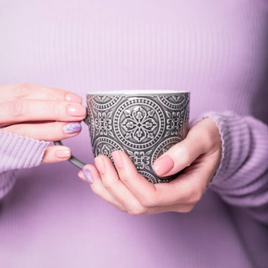 Soluk mor menekşe arka planda gri fincanı tutan güzel kadın elleri. Pembe ojeli, parlak desenli, çıplak manikürlü manikür. Boşluğu kopyala
