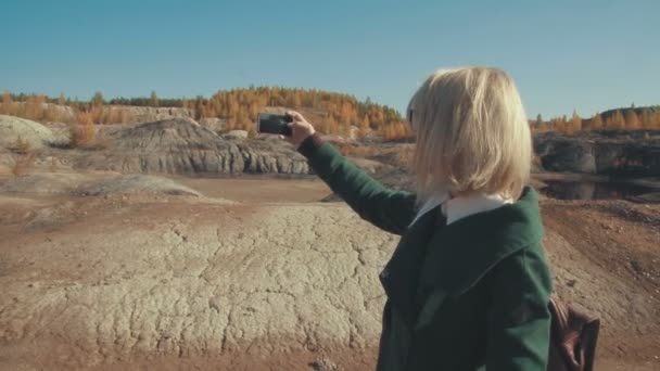Молодая женщина в зеленом пальто фотографирует пустыню со своим смартфоном, любуясь пейзажем — стоковое видео