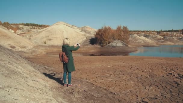 Молода жінка в зеленому пальто фотографує в пустельних пейзажах зі своїм смартфоном, милуючись пейзажем — стокове відео