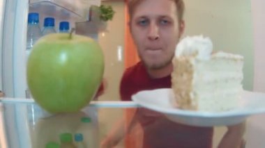 Genç adam bir elma ve pasta arasında seçim yapma buzdolabı açar ve pasta seçer