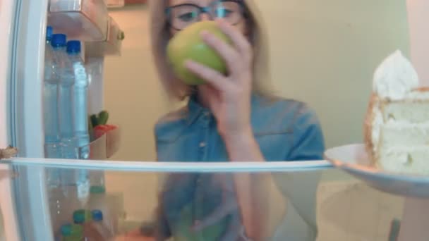 Молода жінка відкриває холодильник, бере свіже яблуко, а потім помічає торт і бере його — стокове відео