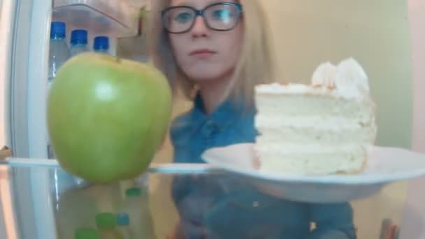 Mujer joven abre el refrigerador sabe pastel, pero toma manzana saludable — Vídeo de stock