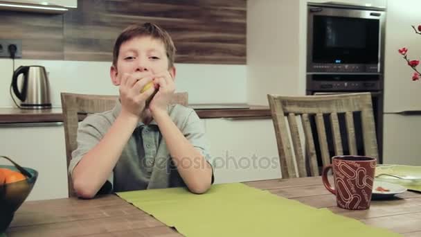 Menino comendo uma maçã sentada na mesa da cozinha — Vídeo de Stock