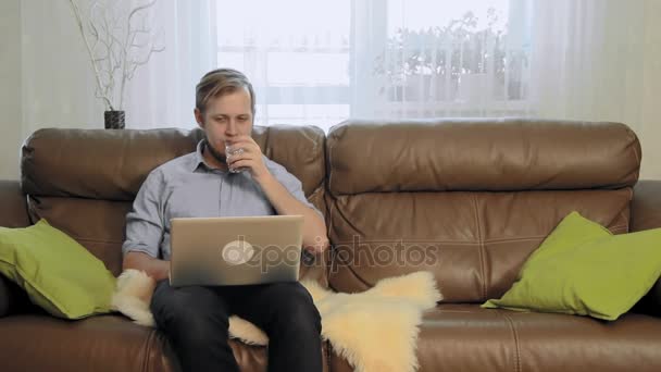 Νεαρός επιχειρηματίας χαλαρώνοντας στο καναπέ και βλέποντας μια ταινία για το φορητό υπολογιστή και πίνοντας κρασί — Αρχείο Βίντεο