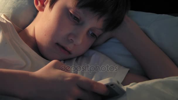 Pojke vila i sin säng på kvällen och med hjälp av sin smartphone — Stockvideo