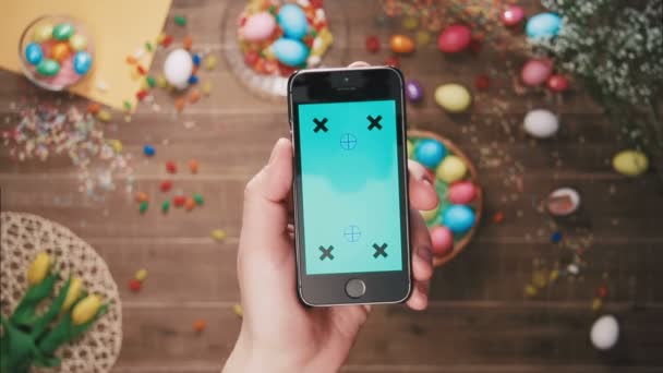 Hand holding phone with green screen in front of table decorated with easter eggs. Top view — Stock Video