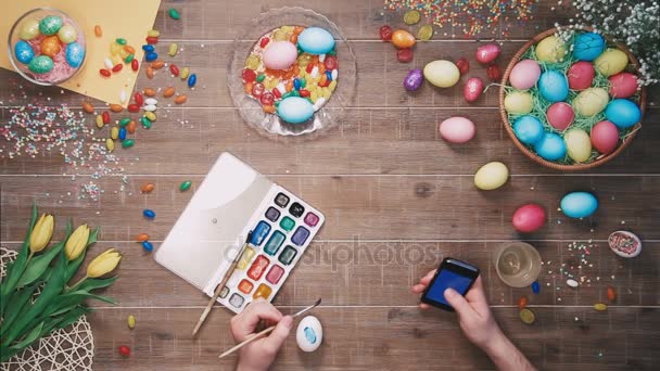 Muž, malování kraslic a pomocí smartphone na stole zdobí velikonoční vajíčka. Pohled shora