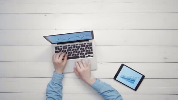 Vista dall'alto mani maschili che lavorano su computer portatile e tablet digitale con grandi grafici di dati che giacciono sulla scrivania bianca da sopra lo spazio di copia — Video Stock