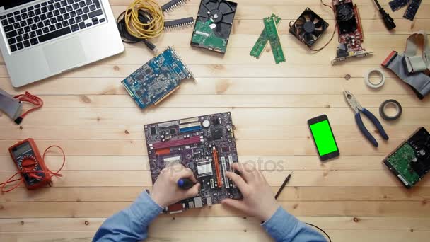 Top view computer technician repairing computer motherboard and smartphone with green screen lying at wooden desk with tools and electronic components — Stock Video