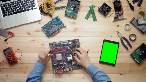 Top view computer technician repairing computer motherboard and digital tablet with green screen lying at wooden desk with tools and electronic components — Stock Video