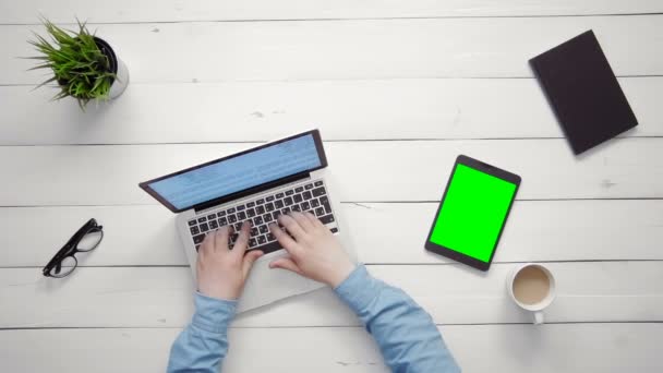 Visão superior mãos masculinas trabalhando no computador portátil e tablet digital com tela verde deitada na mesa branca de cima — Vídeo de Stock