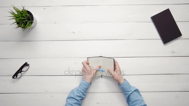 Männliche Hände schreiben in Notizbuch auf weißem Schreibtisch von oben — Stockvideo