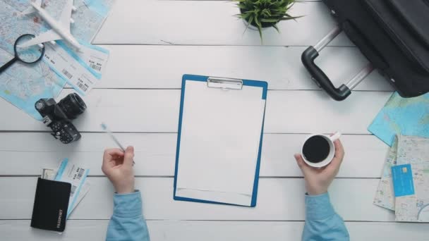 Bovenaanzicht reizigers handen met pen en kopje koffie en Klembord met papier op witte houten bureau — Stockvideo