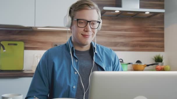 Joven hombre riendo viendo la película en streaming en el ordenador portátil en casa comiendo cereales con auriculares — Vídeo de stock