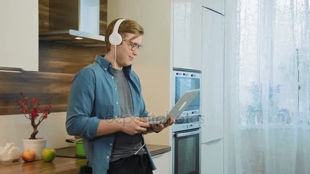 Unga skrattande man titta på strömmande film på laptop äta hemma spannmål med hörlurar — Stockvideo