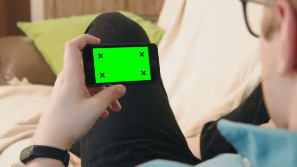 Hombre joven acostado en el sofá mirando el teléfono inteligente con pantalla verde — Vídeo de stock