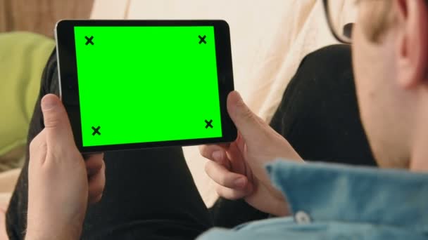 Молодой человек лежит на диване и смотрит на цифровой планшет с зеленым экраном — стоковое видео