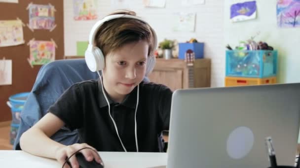 Bonito menino jogando jogo de computador em seu computador portátil usando fones de ouvido — Vídeo de Stock