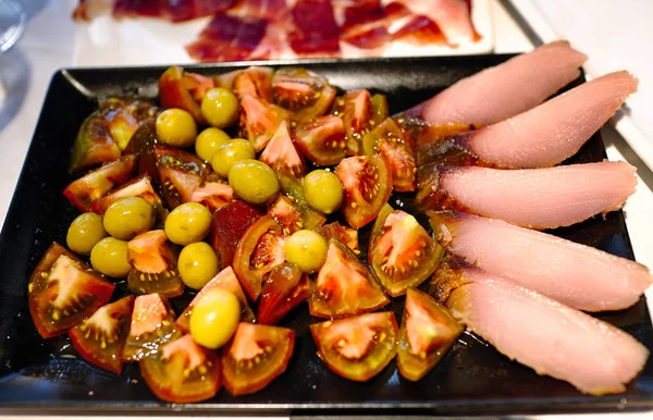 Deliciosa comida, tomates oscuros cortados con aceitunas verdes y rodajas de pescado rojo de mar, en una bandeja negra. Restaurante español —  Fotos de Stock