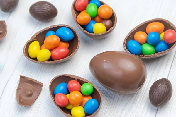 Chocolate Easter eggs and chocolate eggs filled with sweet and colorful candies, on white wood table. Close up.