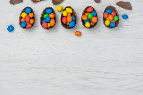 Chocolate Easter eggs and chocolate eggs filled with sweet and colorful candies, on white wood table. Copy space , top view.