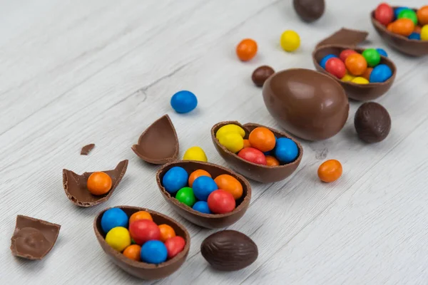 Chocolate Easter eggs and chocolate eggs filled with sweet and colorful candies, on white wood table.