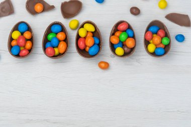 Çikolatalı Paskalya yumurtaları ve çikolatalı yumurtalar beyaz ahşap masada tatlı ve renkli şekerlerle dolu. Boşluğu kopyala, üst görünüm.
