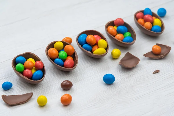 Chocolate Easter eggs and chocolate eggs filled with sweet and colorful candies, on white wood table.