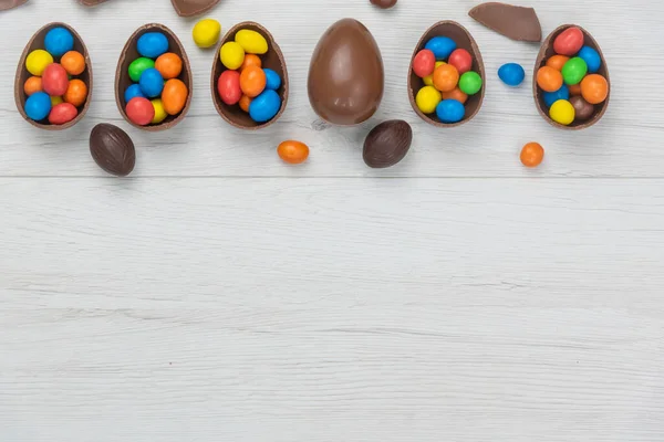 Chocolate Easter eggs and chocolate eggs filled with sweet and colorful candies, on white wood table. Copy space , top view.