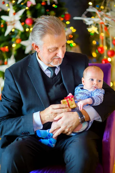 grandfather gives a small grandson a gift and smiles, in the background a festive Christmas tree