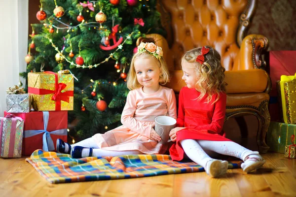 girls sit on the floor near the festive Christmas tree and drink hot te
