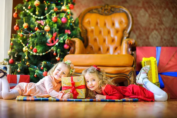 two girls are lying on the floor near gifts and smiling, in the background a festive Christmas tre