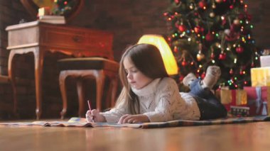 Beyaz örgü kazak güzel küçük kız yerde yatıyor ve şenlikli bir Noel ağacı ve bir sürü hediye Noel Baba'ya bir mektup arka planda yazıyor