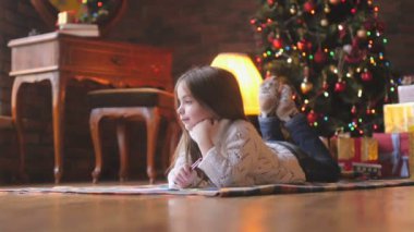 Beyaz örgü kazak güzel küçük kız yerde yatıyor ve şenlikli bir Noel ağacı ve bir sürü hediye Noel Baba'ya bir mektup arka planda yazıyor