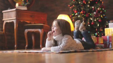 Beyaz örgü kazak, güzel küçük bir kız yere yatıyor, oluşturur ve şenlikli bir Noel ağacı ve hediyeler bir dizi arka planda Noel Baba'ya bir mektup yazar