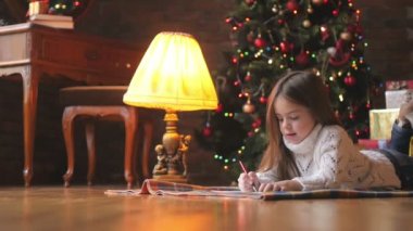 Beyaz örgü kazak güzel küçük kız yerde yatıyor ve şenlikli bir Noel ağacı ve bir sürü hediye Noel Baba'ya bir mektup arka planda yazıyor