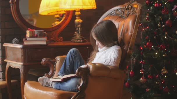美丽的小女孩正在读书 坐在一张靠近节日圣诞树的椅子上 — 图库视频影像