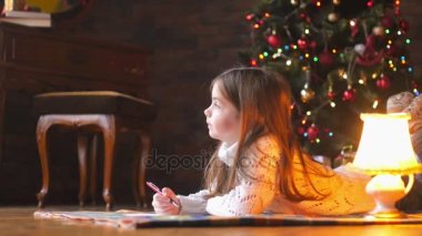 Beyaz örgü kazak, güzel küçük bir kız yere yatıyor, oluşturur ve şenlikli bir Noel ağacı ve hediyeler bir dizi arka planda Noel Baba'ya bir mektup yazar