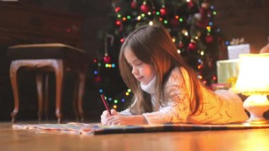 Beyaz örgü kazak güzel küçük kız yerde yatıyor ve şenlikli bir Noel ağacı ve bir sürü hediye Noel Baba'ya bir mektup arka planda yazıyor