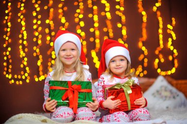 Pijamalı ve Noel Baba şapkalı iki tatlı kız ellerinde hediyelerle yatakta oturuyorlar. Mutlu çocuklar. Noel hediyeleri.